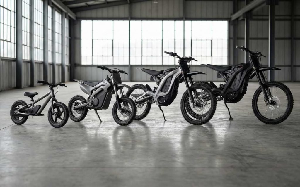 A lineup of four electric dirt bikes progressively increasing in size from a small youth model on the left to full-size adult bikes, including Surron style models, on the right. They are parked on a concrete floor inside a large industrial warehouse with natural light coming through tall windows.