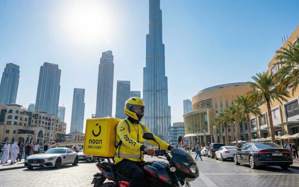 Noon delivery rider in yellow uniform driving a motorcycle in Downtown Dubai with the Burj Khalifa in the background.