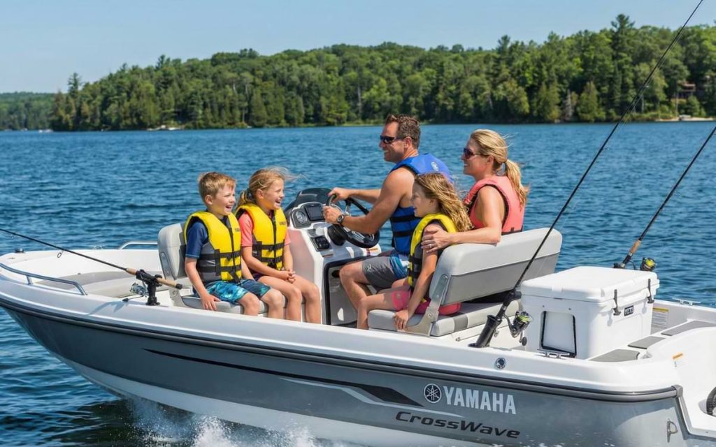 A family of four with two children in life jackets smiles while cruising on a Yamaha CrossWave fishing boat on a sunny lake, with fishing rods mounted on the stern.