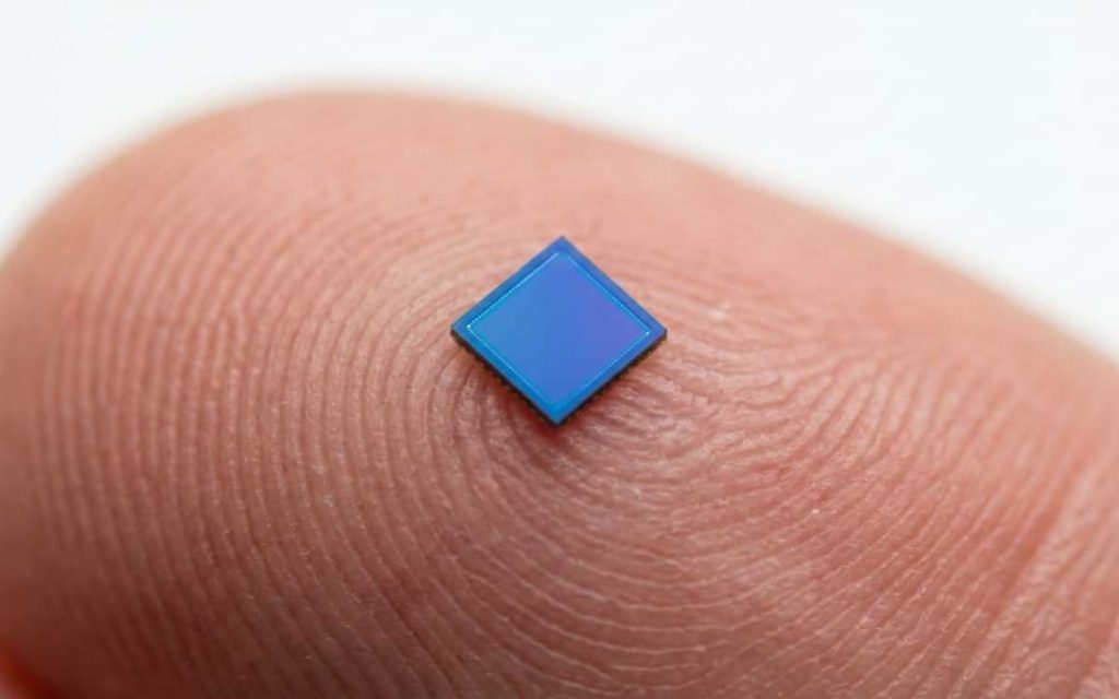 Macro photography close-up of the OmniVision OV6948, the world's smallest image sensor, resting on a human fingertip to demonstrate its grain-of-sand size.